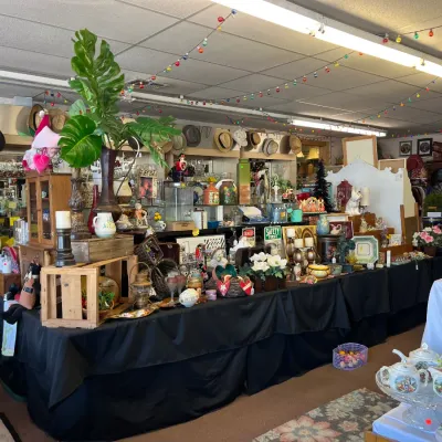 Sale Photo Thumbnail #12: Retail display tables draped in black cloth, featuring an assortment of vintage home decor, artificial plants, teapots, glassware, and hats hung along the wall.