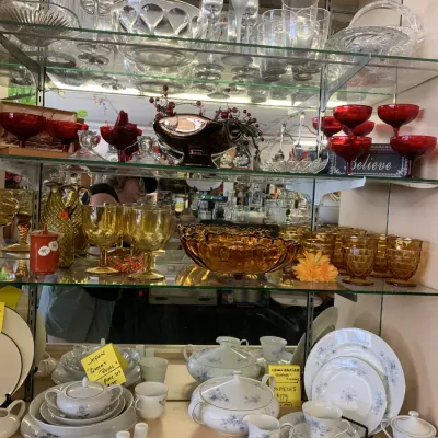 Sale Photo Thumbnail #17: Glass display case filled with vintage glassware, including clear stemware, red dessert dishes, amber pressed glass goblets, and china tea sets.