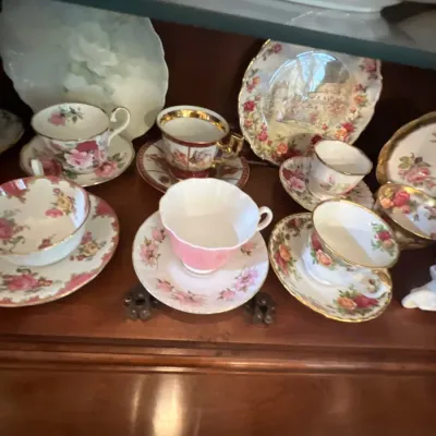 Collection of vintage porcelain teacups and saucers featuring floral patterns and gilded accents displayed inside a hutch.