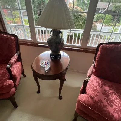 Pair of traditional Queen Anne style armchairs with red patterned upholstery, set beside an oval cherry wood end table topped with a...