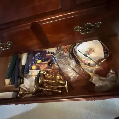 Open wooden dresser drawer containing brass candlesticks, taper candles, floral porcelain tableware, and serving utensils.