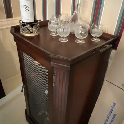Traditional dark wood wine cabinet featuring a mesh front door, fluted corner accents, and a tray-style top displaying glassware and a...
