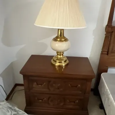 Traditional dark wood two-drawer nightstand with carved detailing, topped with a polished brass and ceramic table lamp with a pleated shade.