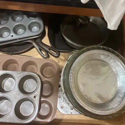 Collection of kitchen bakeware inside a drawer, including metal muffin tins, round cake pans, glass pie dishes, and cast-iron skillets.