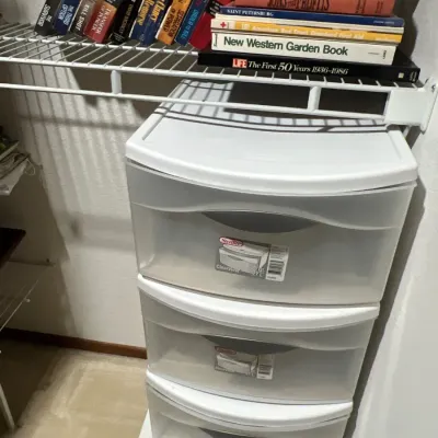 Sterilite white plastic three-drawer storage cart, positioned below a wire shelf holding a collection of paperback and hardcover books.