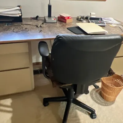 Home office setup featuring a large laminate-top desk, a black faux leather rolling desk chair, a vintage fluorescent desk lamp, and...
