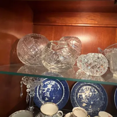 Display cabinet shelf with cut crystal rose bowls and votive holders alongside blue-and-white Transferware plates and porcelain teacups.