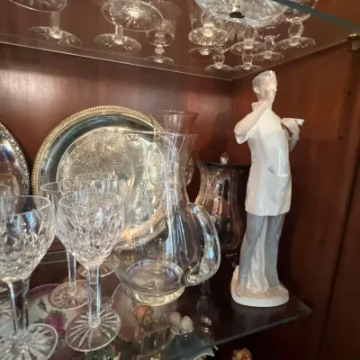 Collection of crystal stemware, silver-toned platters, a clear glass pitcher, and a porcelain Lardro figurine displayed inside a wooden...