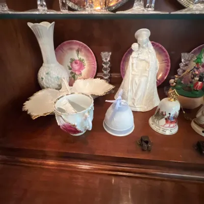Collection of decorative porcelain and glass collectibles displayed inside a wooden cabinet shelf, including a white Madonna statue,...