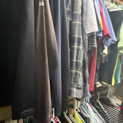 Selection of men's casual clothing hanging in a closet, including plaid button-down shirts, t-shirts, and long-sleeve tops.