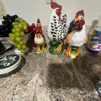 Collection of three hand-painted ceramic rooster figurines displayed on a granite countertop alongside decorative faux fruit.