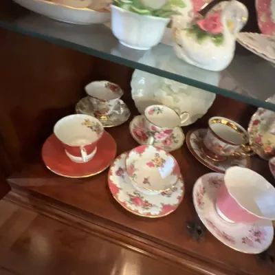 Collection of vintage porcelain teacups and saucers with floral motifs and gilded accents displayed inside a wooden china cabinet.