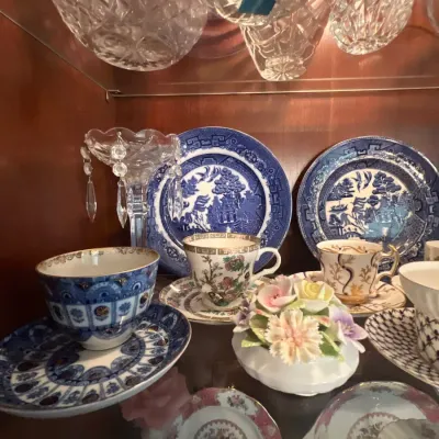 Collection of fine porcelain teacups, saucers, Blue Willow transferware plates, and a porcelain floral figurine displayed inside a glass...