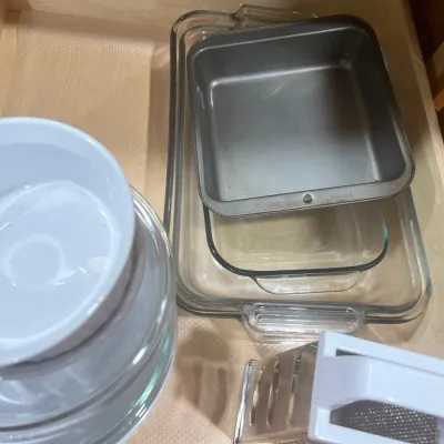 Selection of kitchen cookware and tools stored in a drawer, including clear glass baking dishes, a metal square pan, white mixing bowls,...
