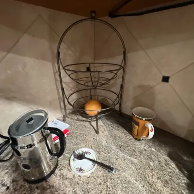 Two-tier metal wire fruit basket shown alongside a stainless steel electric milk frother and kitchen accessories on a granite countertop.