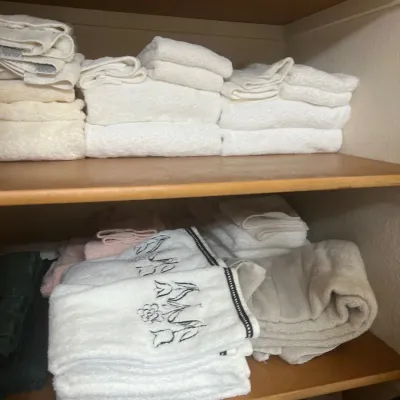 Assorted stacked bath towels and hand towels, including white terrycloth pieces and embroidered designs, arranged on wooden linen closet...