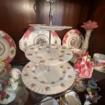 Three-tiered vintage porcelain serving tray with pink floral motifs, surrounded by matching teacups, saucers, and decorative figurines in...