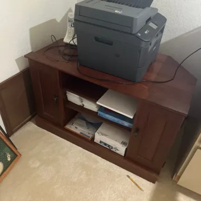 Brother all-in-one laser printer sitting atop a dark wood corner media cabinet or office console with open shelving and side doors.