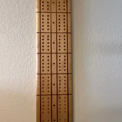 Handcrafted wall-hanging wooden cribbage board featuring inlaid grid lines and top peg storage slots.