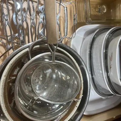 Assorted vintage kitchenware inside a drawer, including metal strainers, colanders, CorningWare dishes, and a wire trivet rack.