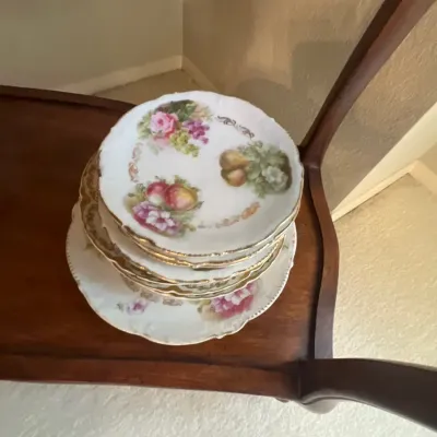 Stack of vintage hand-painted porcelain plates with floral and fruit motifs and gold rim accents, resting on a wooden shelf.
