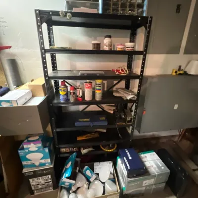 Black metal industrial shelving unit holding various workshop chemicals, lubricants, and hardware, surrounded by boxed lighting supplies.