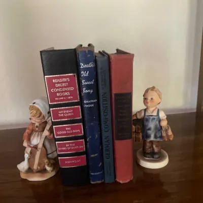Collection of vintage hardcover books flanked by two ceramic Hummles child figurines on a wooden shelf.