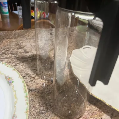 Tall, modern clear glass pitcher with a black plastic handle and lid, sitting on a speckled countertop alongside decorative china platters.