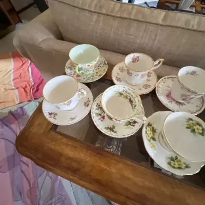 Collection of six fine bone china teacups and matching saucers with various floral patterns, displayed on a glass-top wooden coffee table.