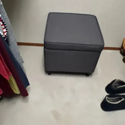 Contemporary dark gray upholstered storage ottoman sitting in a closet floor.