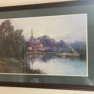 Framed landscape art print featuring a European riverside village with a church spire, housed in a dark wooden frame with a green mat board.