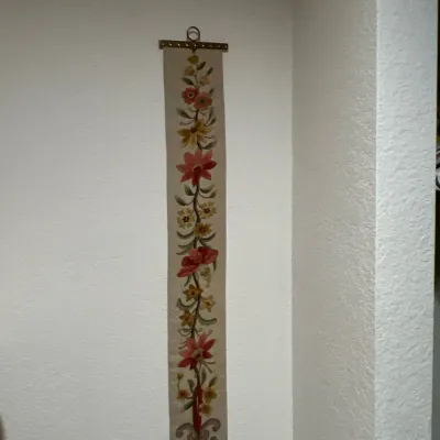 Vintage embroidered bell pull featuring a colorful floral motif and brass hardware hanging on a white wall.