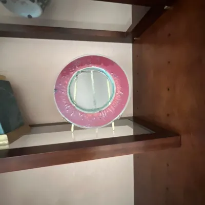 Round decorative glass plate with a textured magenta rim and clear center, displayed on a small brass stand within a wooden shelf.