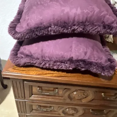 Pair of purple damask throw pillows with brush fringe trim stacked on a vintage wood nightstand.