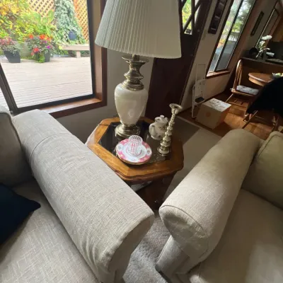 A vintage table lamp with a pleated shade and brass-tone accents sits on a hexagonal wood end table between two beige upholstered...