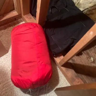 Bright red rolled sleeping bag packed in its matching stuff sack, stored alongside a black garment bag under wooden framing.