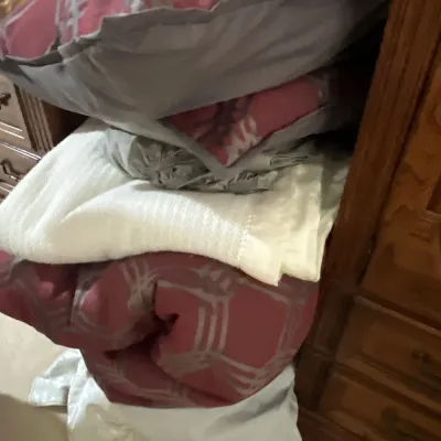 Stack of contemporary bed linens including a dusty rose patterned comforter, decorative throw pillows, sheets, and a white blanket.