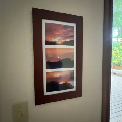 Framed wall art featuring three stacked landscape photographs of a sunset in a dark wood frame with white matting.