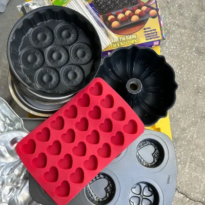 Collection of assorted bakeware including a red silicone heart mold, a non-stick Bundt pan, metal specialty pans, and a boxed Bake Pop...