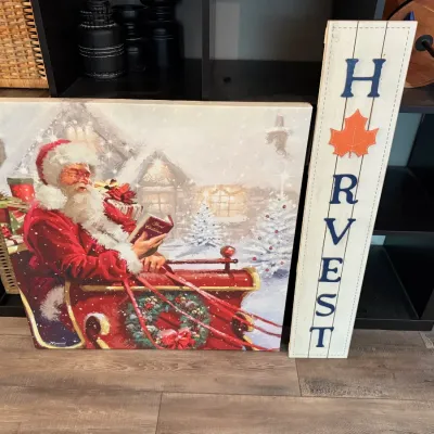 Large canvas print featuring Santa Claus reading in his sleigh, accompanied by a vertical wooden 'HARVEST' porch sign with a...