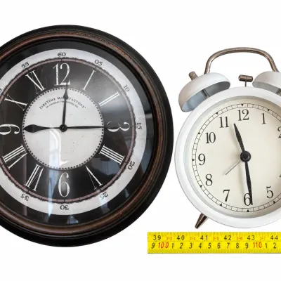 FirsTime Manufactory round wall clock with a black and white dial, shown alongside a vintage-style white twin-bell alarm clock.