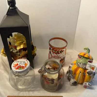 Collection of autumn-themed home decor items, including a black lantern filled with faux flowers, glass jars with festive motifs, and a...