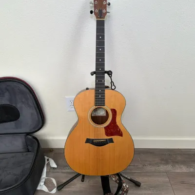 Taylor GS Mini acoustic guitar displayed on a tripod stand, shown alongside its original padded gig bag and guitar strap.