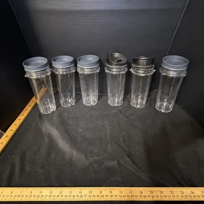 Set of six clear plastic NutriBullet blender cups with dark grey lids, arranged alongside wooden yardsticks for scale.
