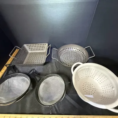 Assorted vintage kitchen colanders and mesh strainers, including metal grilling toppers, fine mesh strainers with black handles, and a...