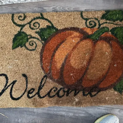 Autumn-themed coir doormat featuring a large painted pumpkin design, green vines, and "Welcome" in black script.