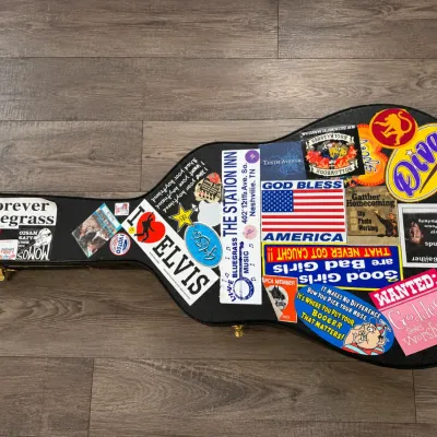 Black hardshell acoustic guitar case heavily decorated with assorted vintage and modern stickers.