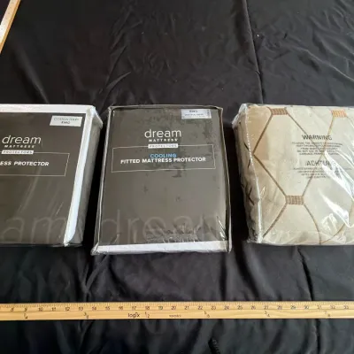 Three packaged king-size bedding accessories, including two Dream Mattress Protectors and a patterned textile item, shown alongside a...