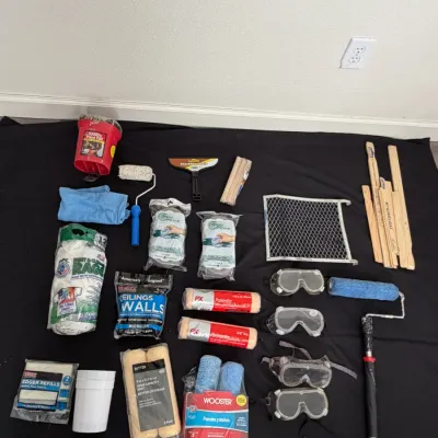 Assortment of painting supplies and safety gear, including Wooster paint roller covers, safety goggles, paint grids, stir sticks, and a...