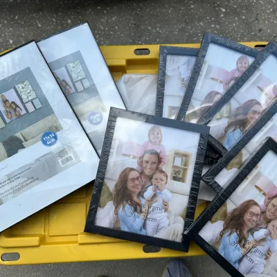 Collection of black Mainstays picture frames, sealed in original plastic packaging and resting on a yellow plastic bin lid.